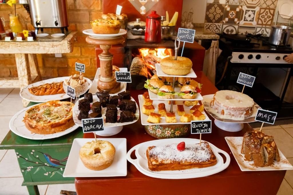Mesa do café com bolos caseiros identificados em plaquinhas, em frente ao fogão a lenha aceso